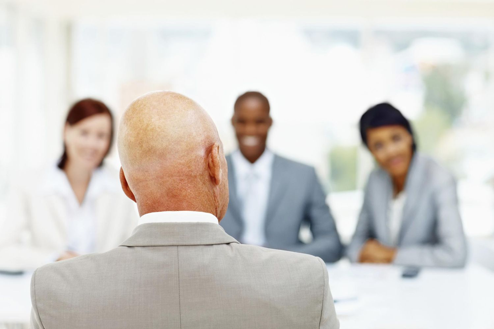 An older man with back to us at an interview panel with 3 interviews who are facing us but are blurred